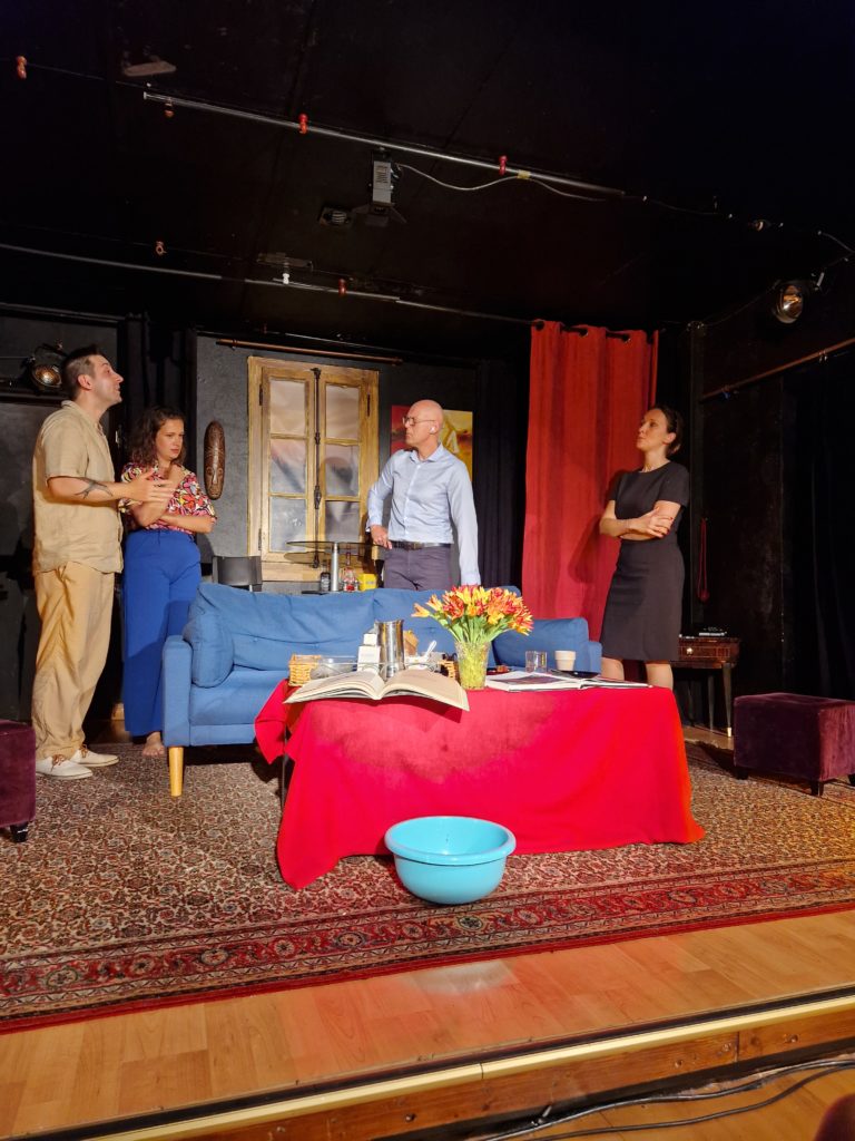 Photo de scène de théâtre sur une scène avce deux homme et deux femme qui parle. Sur la scène il ya un canapé bleu, une table basse avec des fleur et un livre dessus. Derrière eux un rideau rouge et une fentre en bois