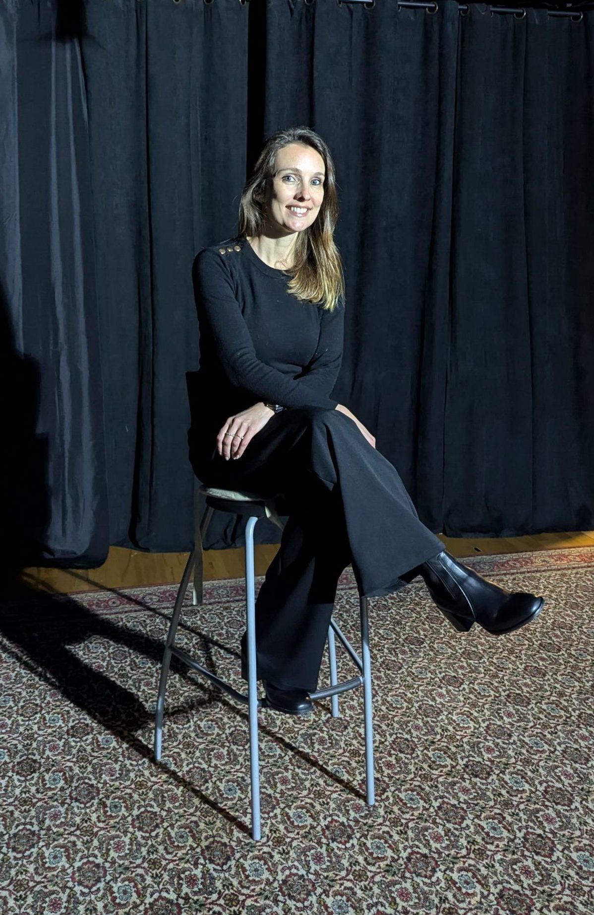 photo d'une femme souriante, habiller en noir assise sur un tabouret, derrière elle un rideau noir de théâtre